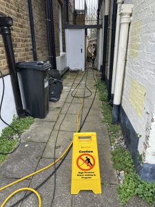 Drain jetting hose through garden with safety signage, South London